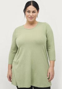 Tunique verte à manches longues avec un col rond, fabriquée en tissu doux, présente un coupe décontractée et des fentes latérales pour plus de facilité de mouvement.