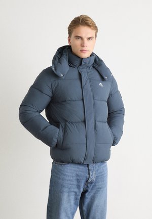 Navy blue puffer jacket with a high collar, hood, snap buttons, and two front pockets; quilted texture and Calvin Klein logo on chest.