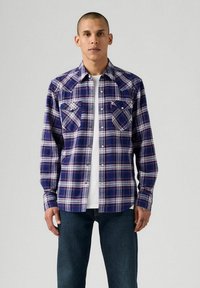Camicia a quadri con bottoni in blu navy, rosso e bianco. Presenta due tasche sul petto e maniche lunghe con polsini a bottone. T-shirt bianca visibile.