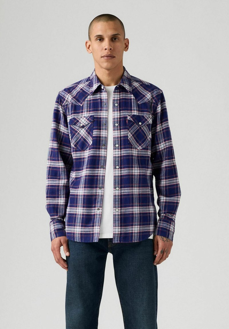 Camicia a quadri con bottoni in blu navy, rosso e bianco. Presenta due tasche sul petto e maniche lunghe con polsini a bottone. T-shirt bianca visibile.