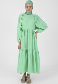Vaaleanvihreä, kerroksellinen maksimekko, jossa on pitkät puff-hihat, kerätty yksityiskohta, ja yhteensopiva huivi. Yhdistetty harmaiden sandaalien kanssa.