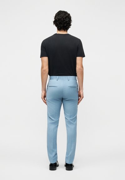 Άνδρας με σγουρά σκούρα μαλλιά στέκεται με την πλάτη, φορώντας μαύρο κοντομάνικο μπλουζάκι, ανοιχτόχρωμο γαλάζιο ραμμένο παντελόνι και μαύρα επίσημα παπούτσια.