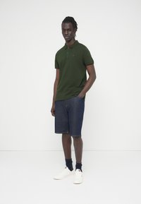 Σκούρο πράσινο πόλο μπλουζάκι, τζιν σορτς και λευκά αθλητικά παπούτσια. Διαθέτει μικρό λογότυπο στο πόλο, με σκουρόχρωμες κάλτσες από ύφασμα. Απλός σχεδιασμός και κλασική εφαρμογή.