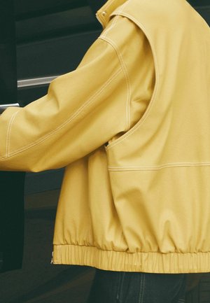 Personne portant une veste jaune avec des coutures blanches ouvrant une porte ou un tiroir de couleur foncée.