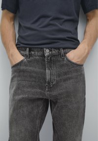 Svarta jeans i denim med en bleknad finish, klassisk femfickdesign och knappstängning. Bärs med en mörk marinblå skjorta, händerna i fickorna.