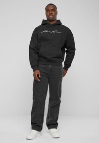Sudadera negra con cordón y bolsillo frontal, con texto blanco. Combinada con jeans gris oscuro y zapatillas blancas. Diseño simple.