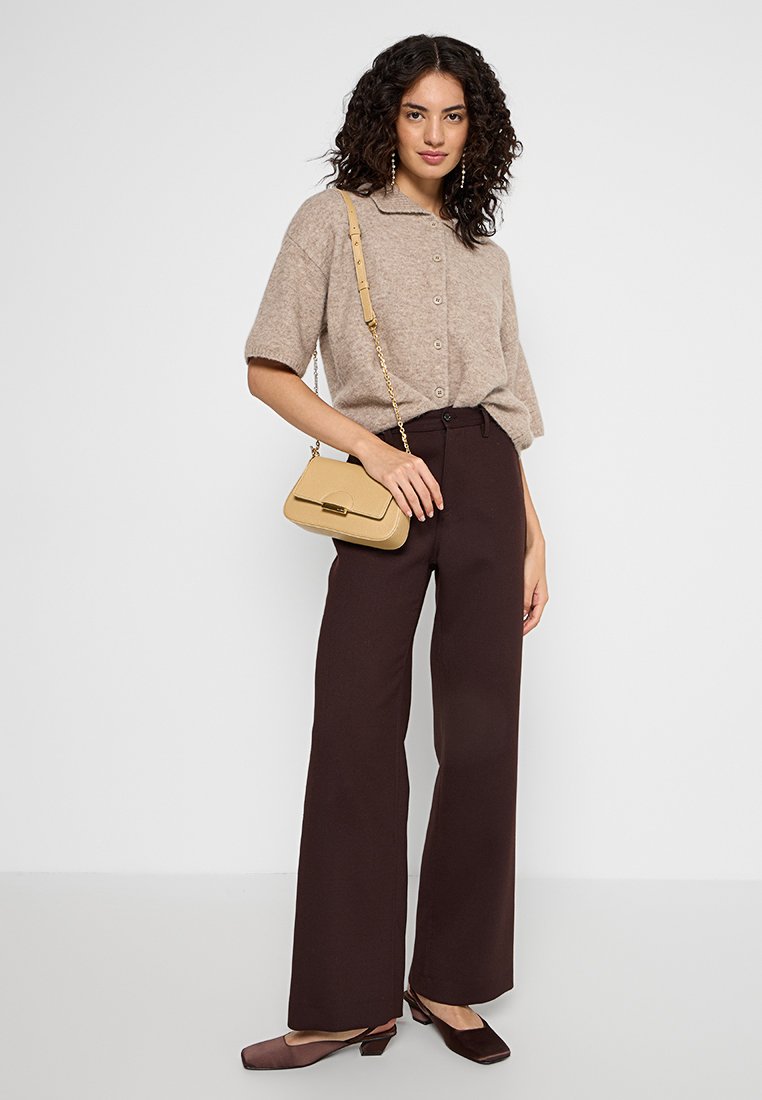 Beżowy sweter z dzianiny z krótkimi rękawami, brązowe spodnie o wysokim stanem i mała torebka typu crossbody w kolorze kremowym. Całość dopełniają brązowe baleriny z ostrymi noskami.