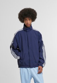 Námornícky modrá bunda s vysokým golierom, biele trojprúžkové akcenty na rukávoch a nášivka s logom. Má voľný strih a hladký povrch.