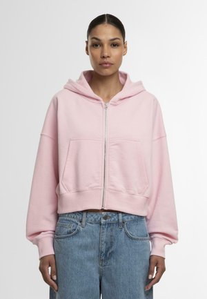 Rózsaszín, rövidített kapucnis pulóver, elülső cipzárral, nagy zsebekkel és oversized ujjakkal, puha anyagból készült. Magas derekú kék farmerral párosítva.
