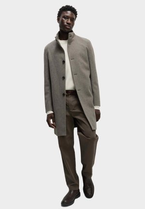 Abrigo de lana gris con cuello alto y cierre de botones, combinado con un suéter crema y pantalones marrones. Botas marrones completan el look.