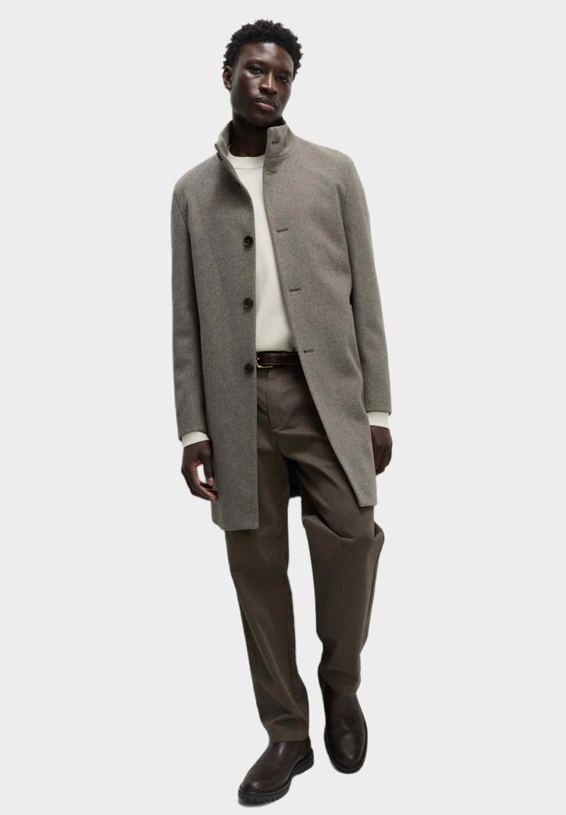 Abrigo de lana gris con cuello alto y cierre de botones, combinado con un suéter crema y pantalones marrones. Botas marrones completan el look.