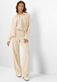 Beige geribbelde cardigan, witte blouse met ruches, beige broek met riem en witte schoenen. De outfit heeft een relaxte pasvorm en een gestructureerde stof.