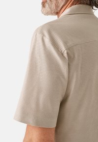 Camicetta a maniche corte in un leggero beige, caratterizzata da un design tessuto testurizzato. Include un colletto e lievi dettagli di cucitura.