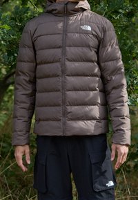 Hnědá péřová bunda se zapínáním na zip, horizontálními prošívanými pruhy a kapucí. Logo na levé straně hrudi. Nošena s černými cargo kalhotami.