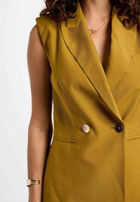 Chaleco de color mostaza de corte doble, con cortes en los hombros, detalles de botones dorados y bolsillos laterales, confeccionado en una tela suave.