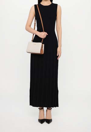 Donna con abito nero lungo senza maniche che tiene una borsa a tracolla beige con monogramma, indossa decolleté nere con cinturino alla caviglia e punta appuntita, in piedi su sfondo bianco.