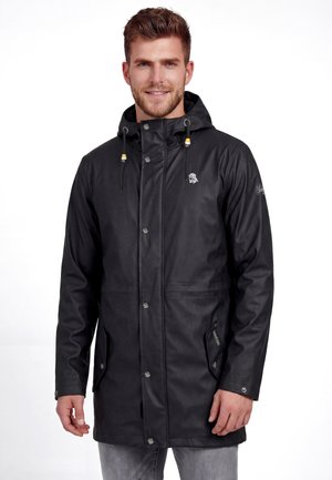 Férfi fekete, vízálló, kapucnis kabátban, gombokkal zárt eleje és zsebek láthatók, fehér, egyszínű háttér előtt áll.