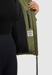 Olijfgroene jas met rits, zwarte voering en stoffen label met tekst. Gladde textuur en verstelbare taille details.