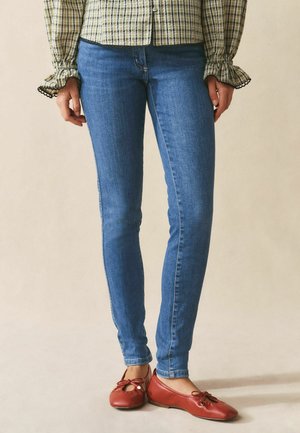 Femeie purtând blugi skinny albaștri, bluză cu mâneci lungi în carouri și balerini roșii, stând pe un fundal neutru.