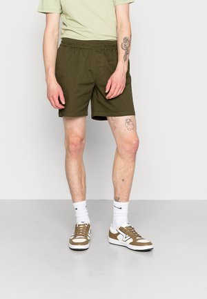 Olijfgroene shorts met een elastische tailleband, gecombineerd met wit en bruine sneakers. Model staat op een grijze vloer en toont been tatoeages.