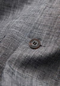 Gros plan sur un tissu gris texturé avec un bouton noir à quatre trous marqué "OMBRE" cousu sur le matériau.