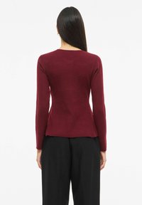 Burgundia pikkade varrukatega pealis, millel on keha järgnev siluett, tekstuuriga soonikkoes ja lehviva äärega, kombineeritud lahtiste mustade pükstega.