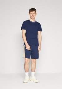 T-shirt bleu marine avec un col côtelé blanc, associé à un short bleu marine avec un panneau latéral beige et un détail de logo, complété par des baskets blanches.
