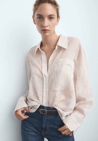 Blusa a righe in crema chiaro e corallo, maniche lunghe, chiusura con bottoni, con tasca. Abbinata a jeans blu scuro e una cintura marrone.