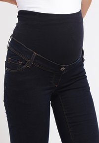 Jeans de maternidade com um corpo de denim azul-escuro e uma cinta elástica preta. As características incluem bolsos frontais e costura em contraste.
