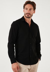 Camisa negra de manga larga con botones y cuello, textura suave, tapeta delantera y botones negros. Combinada con pantalones de color claro.