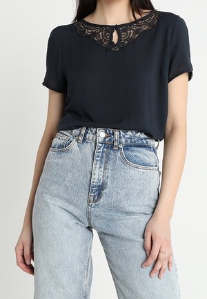 Kvinde iført en sort top med korte ærmer og blonder ved halsudskæringen samt lyseblå jeans med høj talje, stående foran en ensfarvet baggrund.