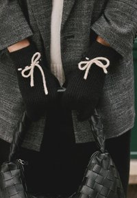 Schwarze gestrickte Handschuhe mit beigen Bandakzenten, getragen von einer Person, die eine gewebte schwarze Handtasche hält, kombiniert mit einem grauen Karomantel.