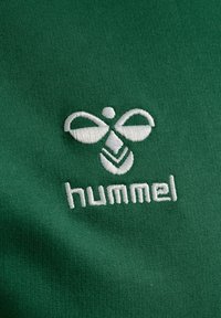 Groene stof met een gladde textuur met een wit geborduurd logo dat lijkt op een gestileerde figuur. De merknaam "hummel" is ook geborduurd.