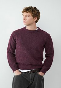 Burgunderfarbener Wollpullover mit Rundhalsausschnitt und Rippbündchen; getragen über einem weißen Hemd, kombiniert mit dunklen Jeans und einem braunen Gürtel.