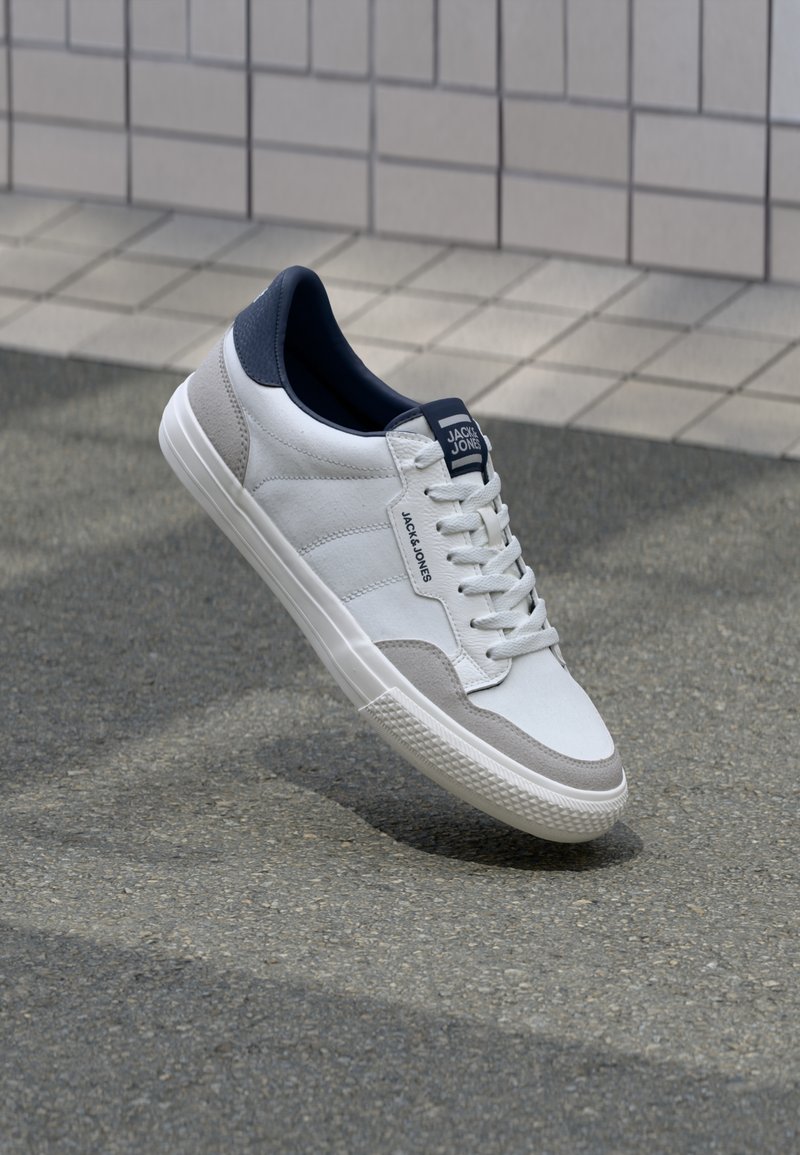 Sneaker bianca con dettagli grigi e blu navy, caratterizzata da una parte superiore in pelle liscia e scamosciata, chiusura con lacci e suola in gomma testurizzata.
