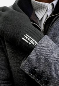 Guante de punto negro con textura acanalada, que presenta una marca blanca, superpuesto a un abrigo gris de espiga con botones oscuros y una camisa a rayas.