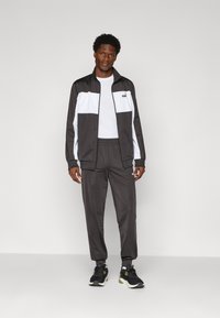 Man wearing black and white Puma tracksuit with black sneakers, standing against plain light gray background.