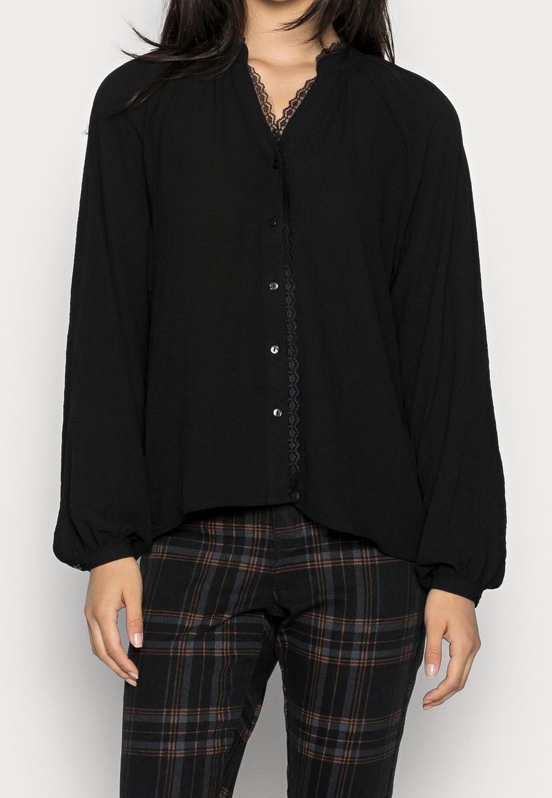Femme portant un chemisier noir boutonné avec bordure en dentelle et manches longues bouffantes, associé à un pantalon à carreaux noirs et marron.
