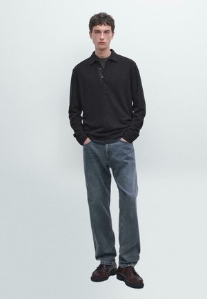 Young man standing with hands in pockets wearing black long-sleeve polo, gray jeans, and dark leather shoes against plain background.