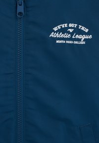 Tissu bleu avec une fermeture éclair et un texte brodé blanc indiquant "We've got this Athletic League North Road College" sur le côté droit.