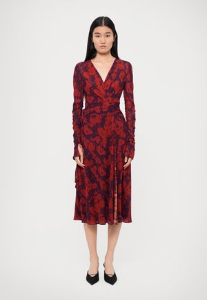 Langsleeve, wikkeljurk in het rood met een donker patroon, met een diepe V-halslijn en een vloeiende rok. Gecombineerd met puntige zwarte hielen.