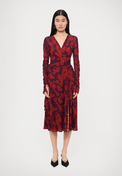 Robe enveloppante à manches longues en rouge avec un motif sombre, présentant un décolleté en V profond et une jupe fluide. Associée à des talons aiguilles noirs.