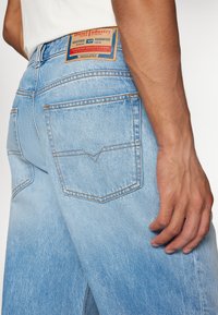 Jeans de mezclilla de color azul claro con un ligero desgaste, que presentan una etiqueta de cuero marrón en la cintura y dos bolsillos traseros con un diseño cosido.