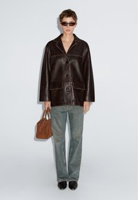 Bruine leren jas met gedetailleerd stiksel, oversized pasvorm, knopen aan de voorkant en twee zakken. Gecombineerd met lichtgewassen spijkerbroek en een kleine bruine handtas.