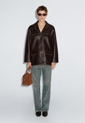 Hnědá kožená bunda s detailním prošíváním, volným oversized střihem, předními knoflíky a dvěma kapsami. Kombinována se světlými džínami a malou hnědou kabelkou.