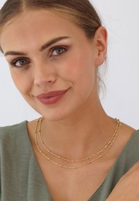 Goldfarbene Halskette mit zarten Ketten und kleinen runden Akzenten, getragen über einem sanften grünen Kleidungsstück. Die glatte Textur verleiht Eleganz.