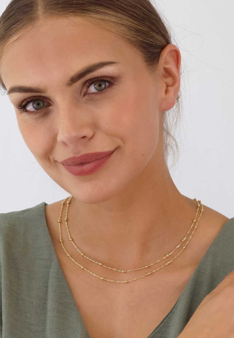 Goldfarbene Halskette mit zarten Ketten und kleinen runden Akzenten, getragen über einem sanften grünen Kleidungsstück. Die glatte Textur verleiht Eleganz.