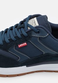Granatowe buty sportowe wykonane z tkaniny i zamszu, z gumową podeszwą, kontrastowym szwem oraz czerwoną metką z logo Levi's na cholewce.