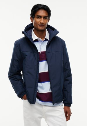 Homme portant une veste zippée bleu marine sur un polo blanc, bordeaux et bleu rayé avec un pantalon blanc, debout, une main dans la poche.