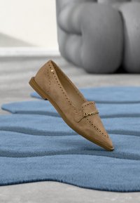 Chaussure plate en daim camel avec des détails perforés et une boucle dorée, posée sur un tapis bleu texturé, avec un canapé gris capitonné en arrière-plan.